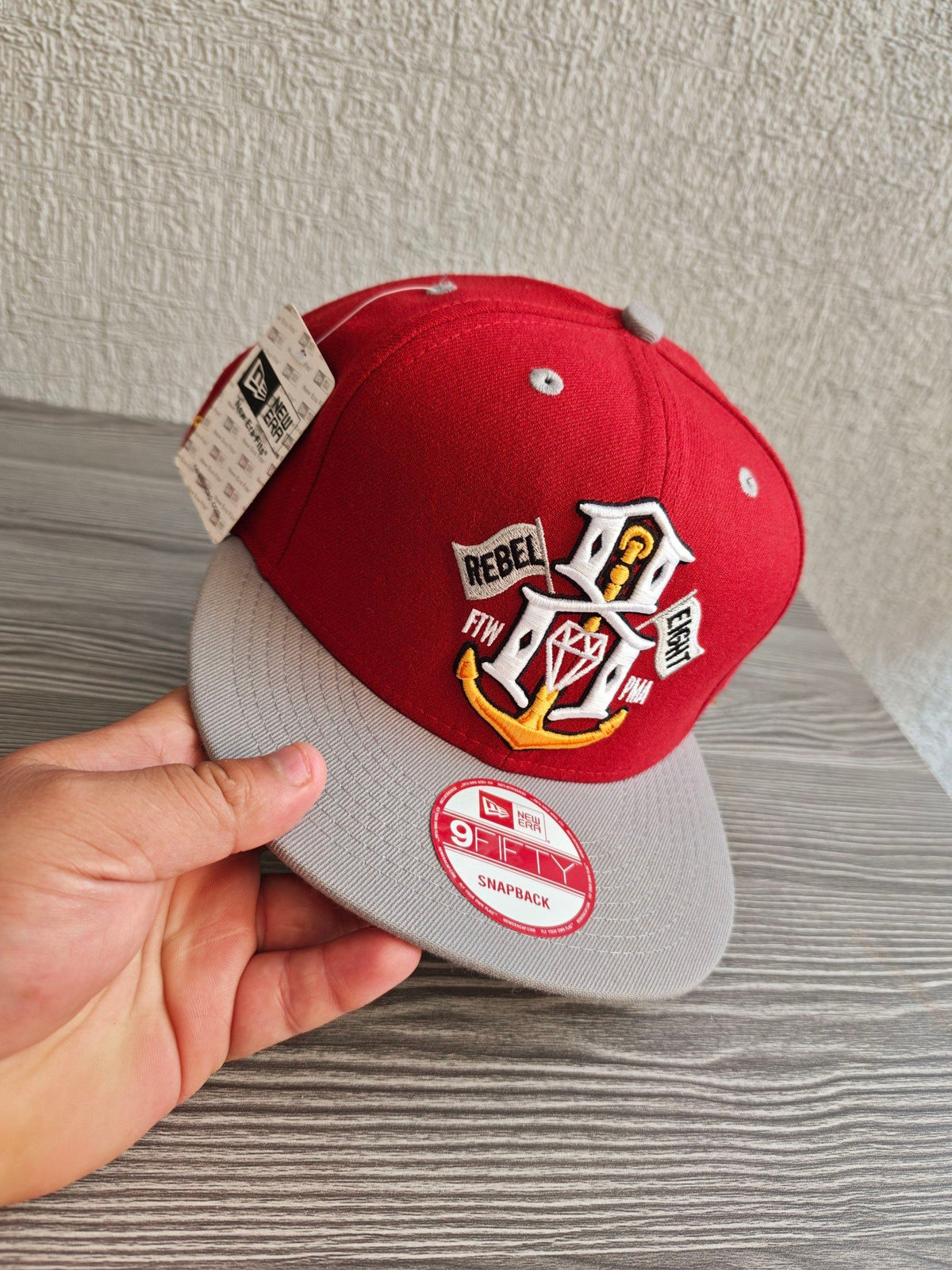 New Era grey and red snapback cap with embroidered "REBEL" anchor design and adjustable strap.