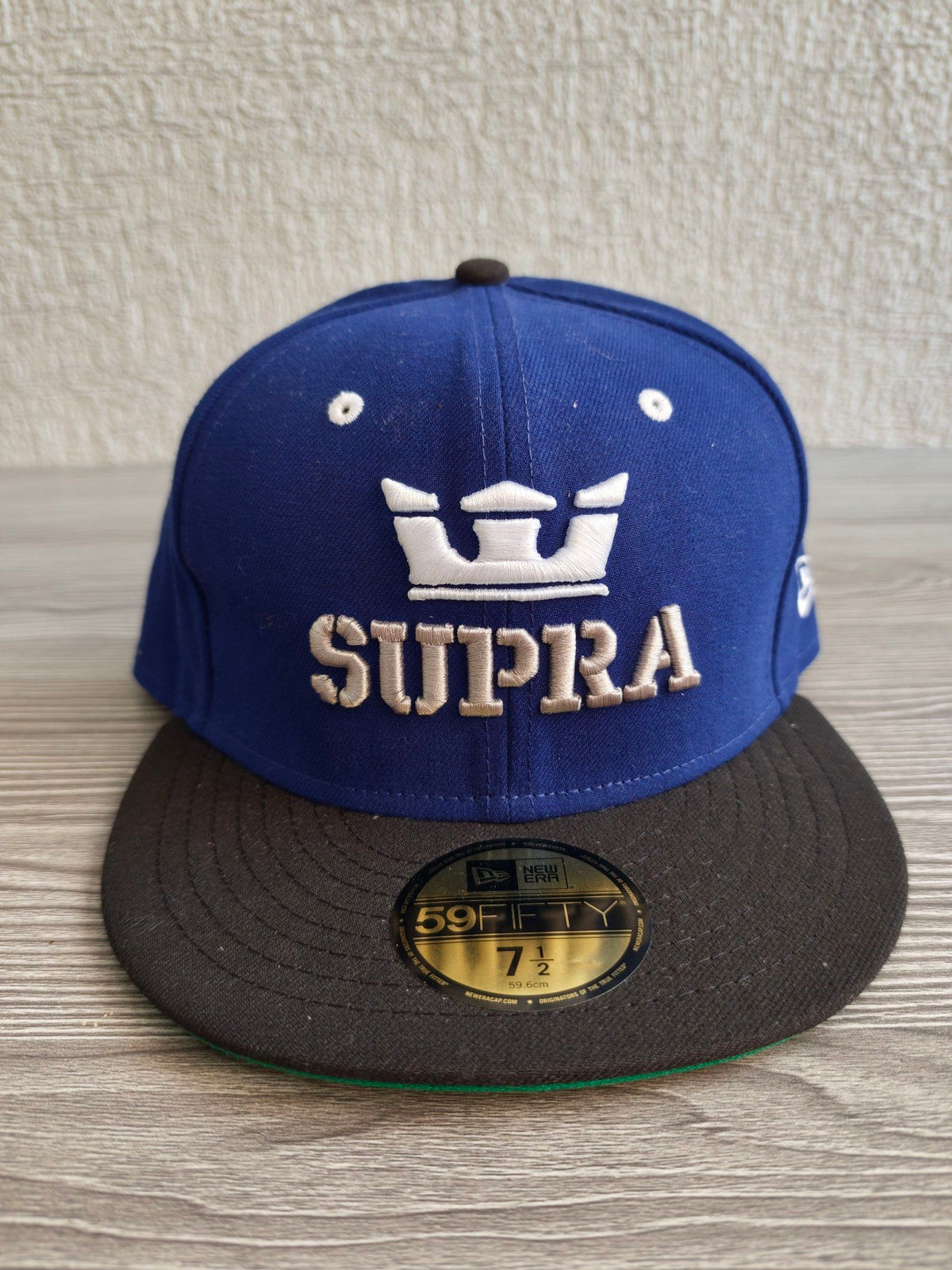 New Era blue and black fitted hat with white embroidered Supra logo and green underside.