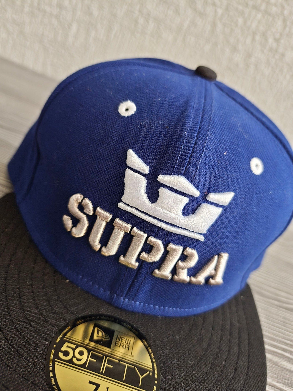 New Era blue and black fitted hat with white embroidered Supra logo and green underside.