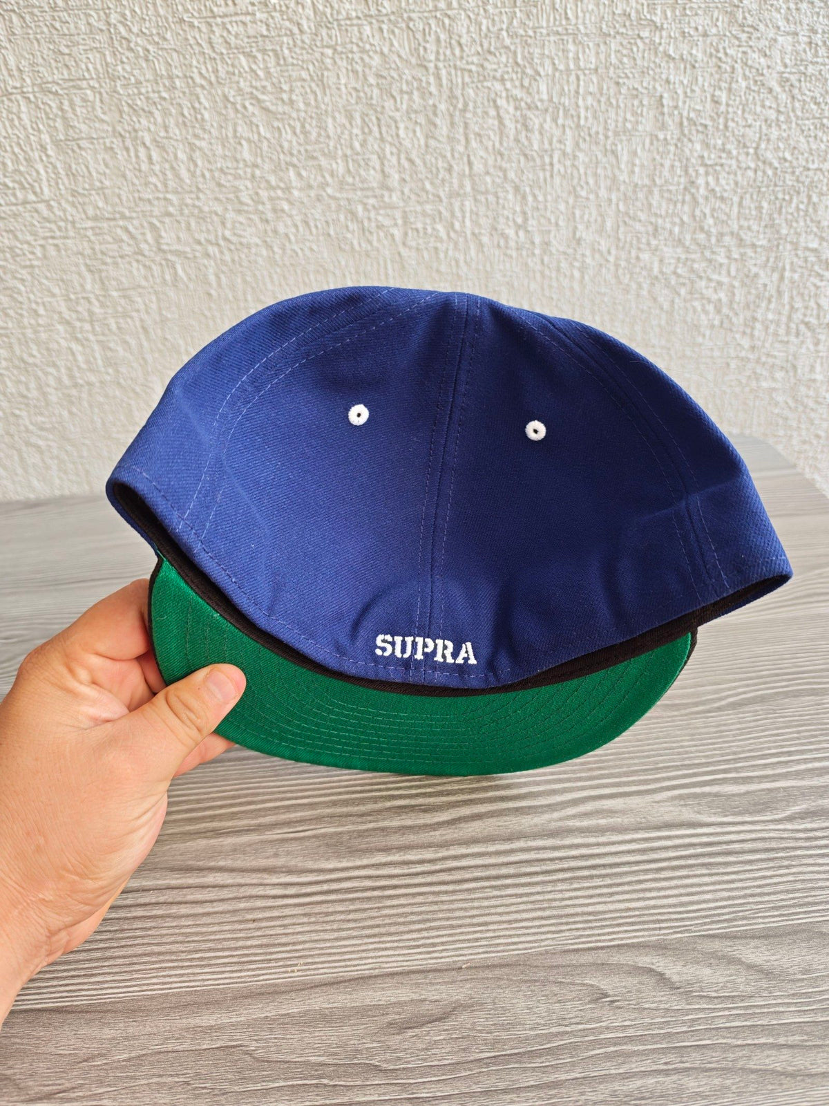 New Era blue and black fitted hat with white embroidered Supra logo and green underside.