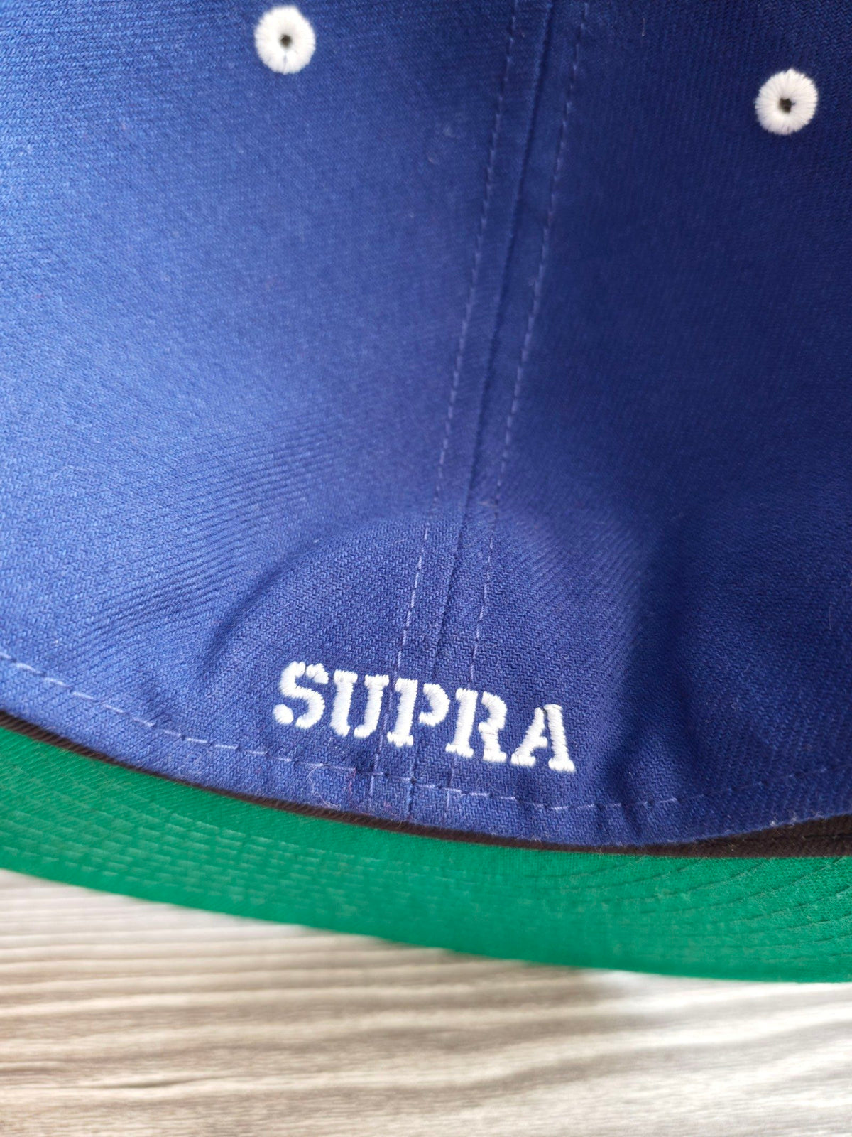 New Era blue and black fitted hat with white embroidered Supra logo and green underside.